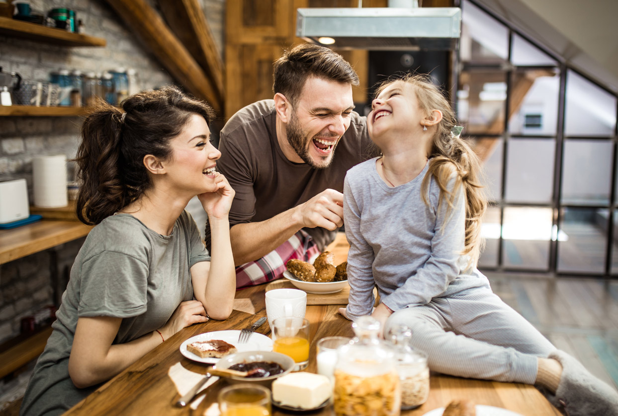 Lachende Familie beim Frühstück: Mutter, Vater und Tochter sitzen an einem Holztisch in einer gemütlichen Küche und genießen gemeinsam das Frühstück.