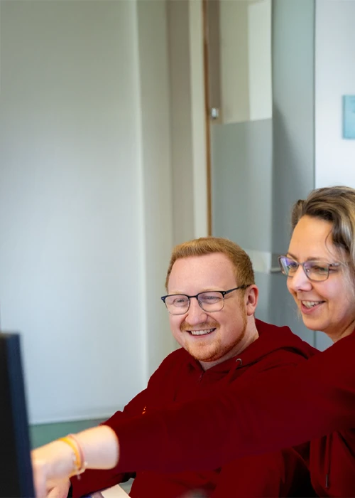 Drei BSH Mitarbeitende in roten Oberteilen sitzen gemeinsam vor einem Computerbildschirm und lachen, während eine Person auf den Monitor zeigt.