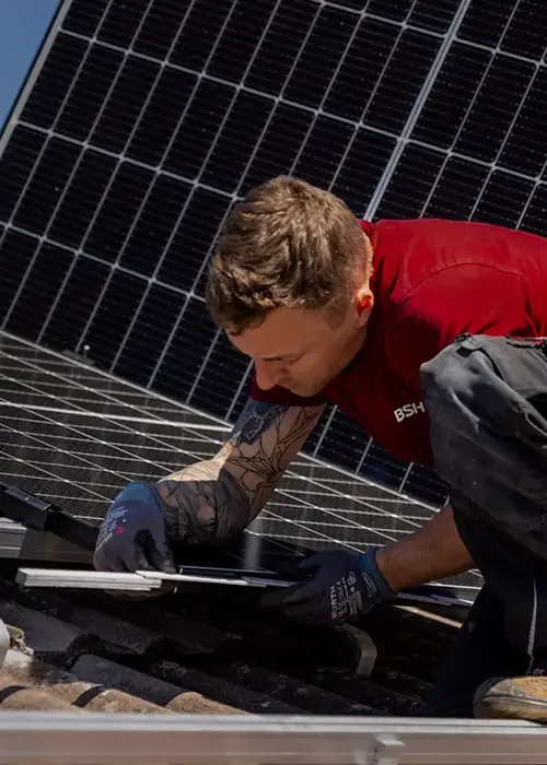 BSH Techniker in rotem Shirt montiert eine Photovoltaikanlage auf einem Ziegeldach bei Sonnenschein.