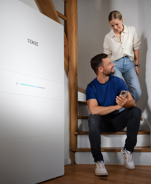 Ein Mann sitzt auf einer Treppe und steuert über sein Smartphone einen SENEC-Stromspeicher, während eine Frau lächelnd neben ihm steht – moderne Energieunabhängigkeit im eigenen Zuhause.