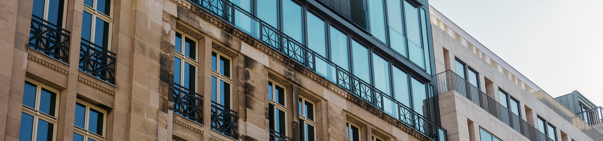 Moderne Gebäudefassade mit klaren Linien, großen Glasflächen und Balkonen, kombiniert mit einer hellen Steinfassade. Mischung aus historischer Architektur und zeitgenössischem Design.
