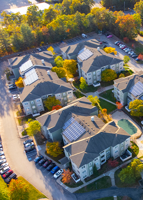 Luftaufnahme einer Wohnanlage mit mehreren Mehrfamilienhäusern, auf deren Dächern Solaranlagen installiert sind, umgeben von Straßen, parkenden Autos und herbstlich gefärbten Bäumen.