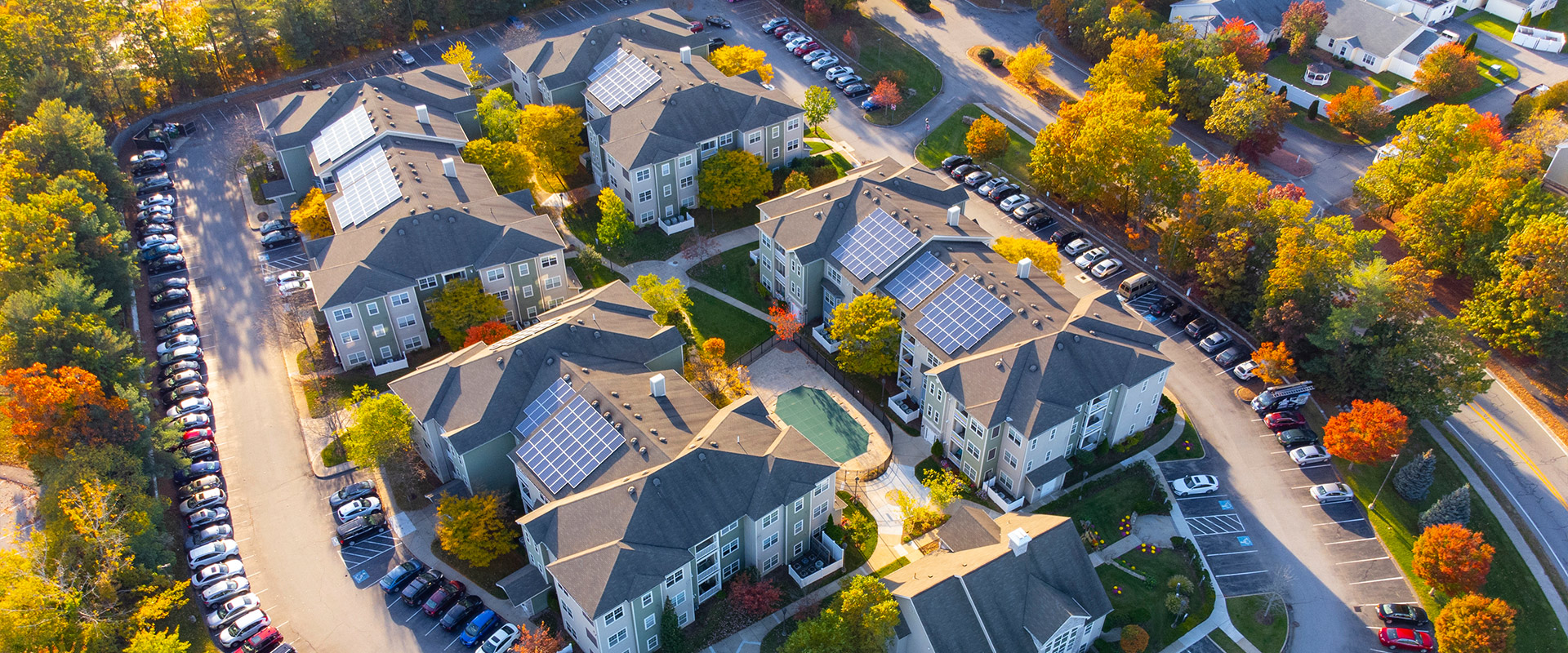Luftaufnahme einer Wohnanlage mit mehreren Mehrfamilienhäusern, auf deren Dächern Solaranlagen installiert sind, umgeben von Straßen, parkenden Autos und herbstlich gefärbten Bäumen.