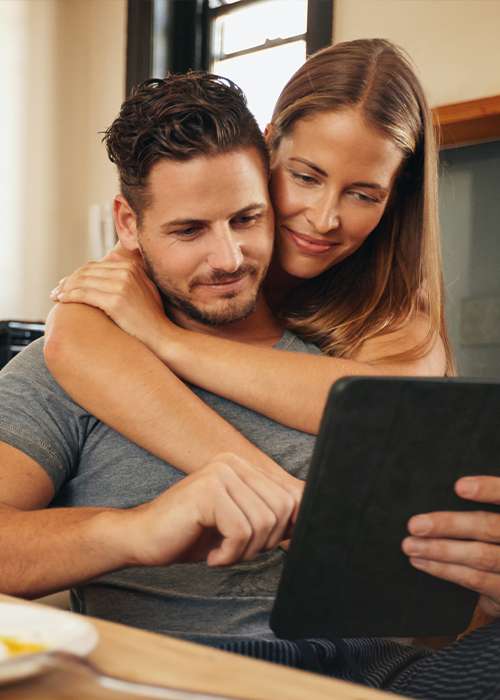 Ein Paar sitzt in einer hellen Küche, die Frau legt dem Mann die Arme um die Schultern, während beide gemeinsam auf ein Tablet schauen.