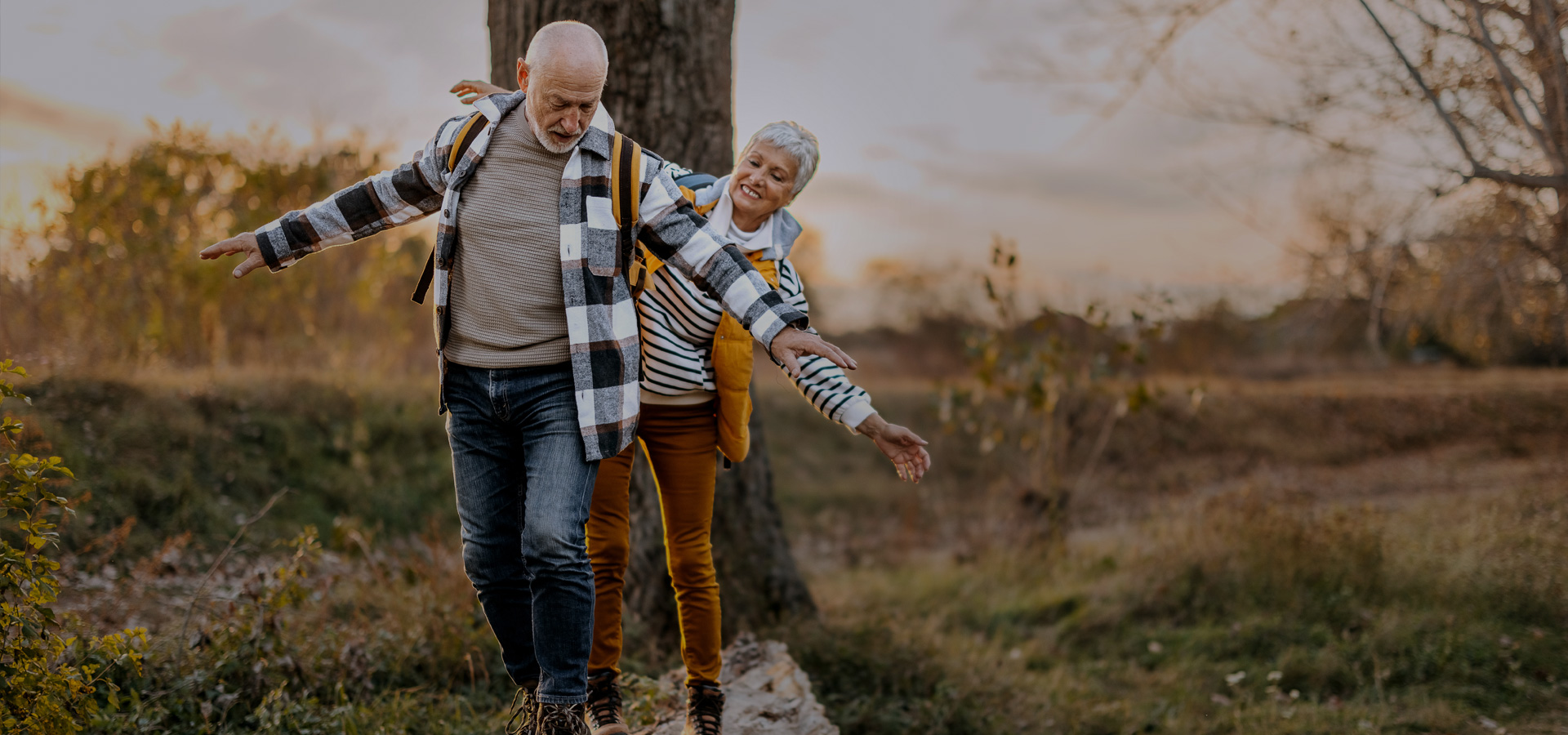 Älteres Paar balanciert lachend auf einem Baumstamm bei einem Spaziergang in herbstlicher Natur.