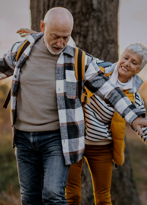 Älteres Paar balanciert lachend auf einem Baumstamm bei einem Spaziergang in herbstlicher Natur.