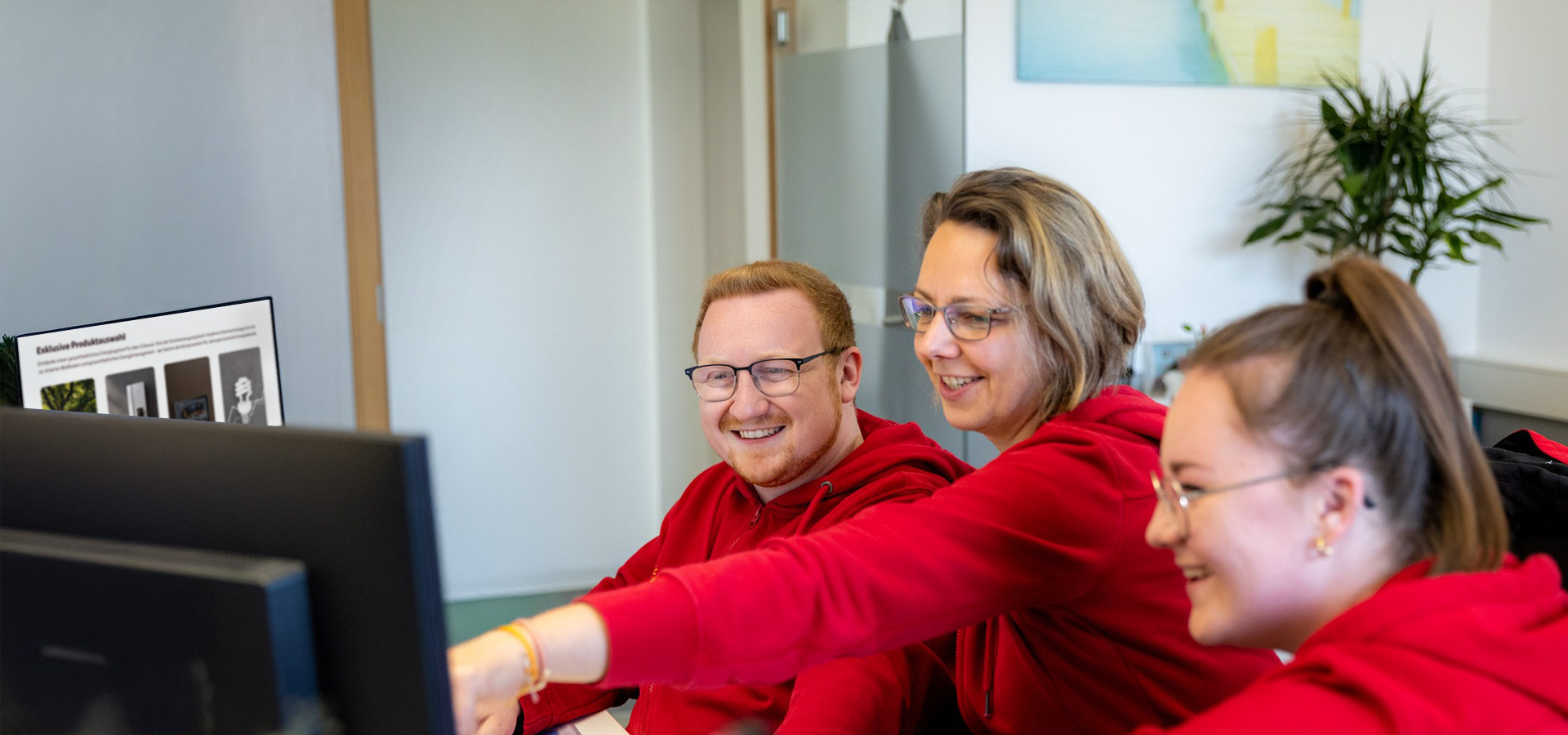 Drei BSH Mitarbeitende in roten Oberteilen sitzen gemeinsam vor einem Computerbildschirm und lachen, während eine Person auf den Monitor zeigt.