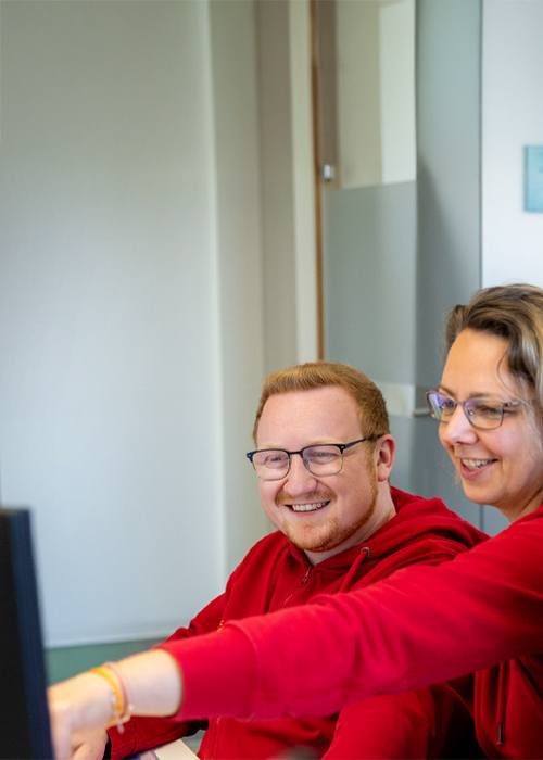 Drei BSH Mitarbeitende in roten Oberteilen sitzen gemeinsam vor einem Computerbildschirm und lachen, während eine Person auf den Monitor zeigt.