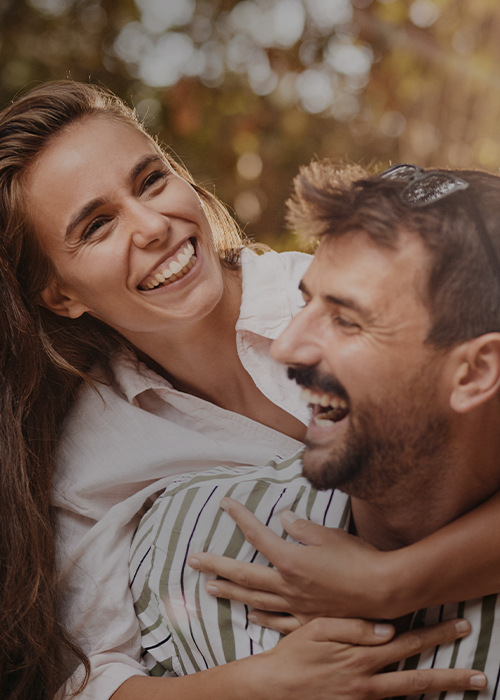 Lächelndes Paar im Freien, der Mann trägt die Frau auf den Schultern, beide genießen den Moment in der Sonne.