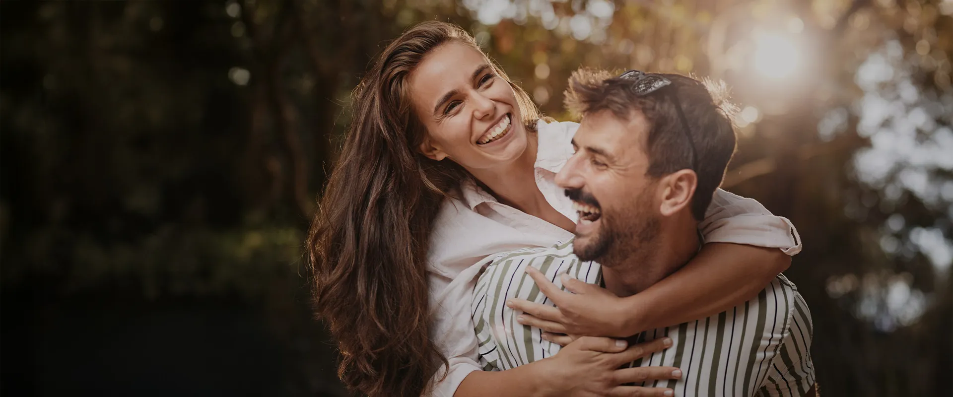 Lächelndes Paar im Freien, der Mann trägt die Frau auf den Schultern, beide genießen den Moment in der Sonne.