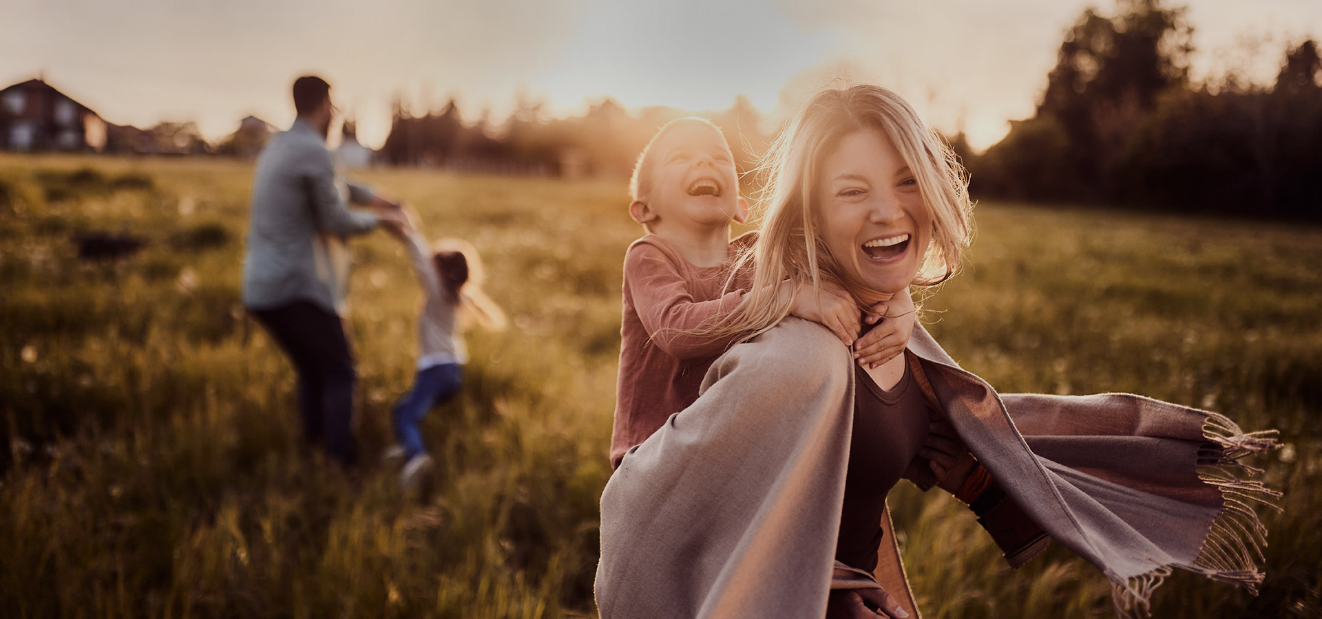 Fröhliche Familie spielt im Sonnenuntergang auf einer Wiese – Mutter trägt lachendes Kind auf dem Rücken, im Hintergrund tanzt der Vater mit einem weiteren Kind.