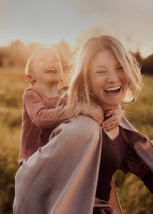 Fröhliche Familie spielt im Sonnenuntergang auf einer Wiese – Mutter trägt lachendes Kind auf dem Rücken, im Hintergrund tanzt der Vater mit einem weiteren Kind.