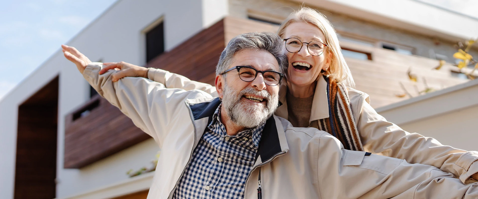 Fröhliches älteres Paar mit ausgebreiteten Armen steht vor einem modernen Haus und lächelt in die Kamera.