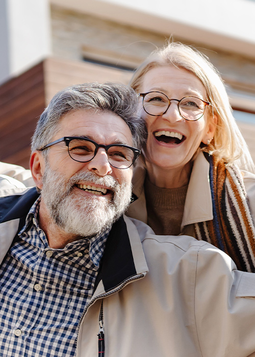 Fröhliches älteres Paar mit ausgebreiteten Armen steht vor einem modernen Haus und lächelt in die Kamera.
