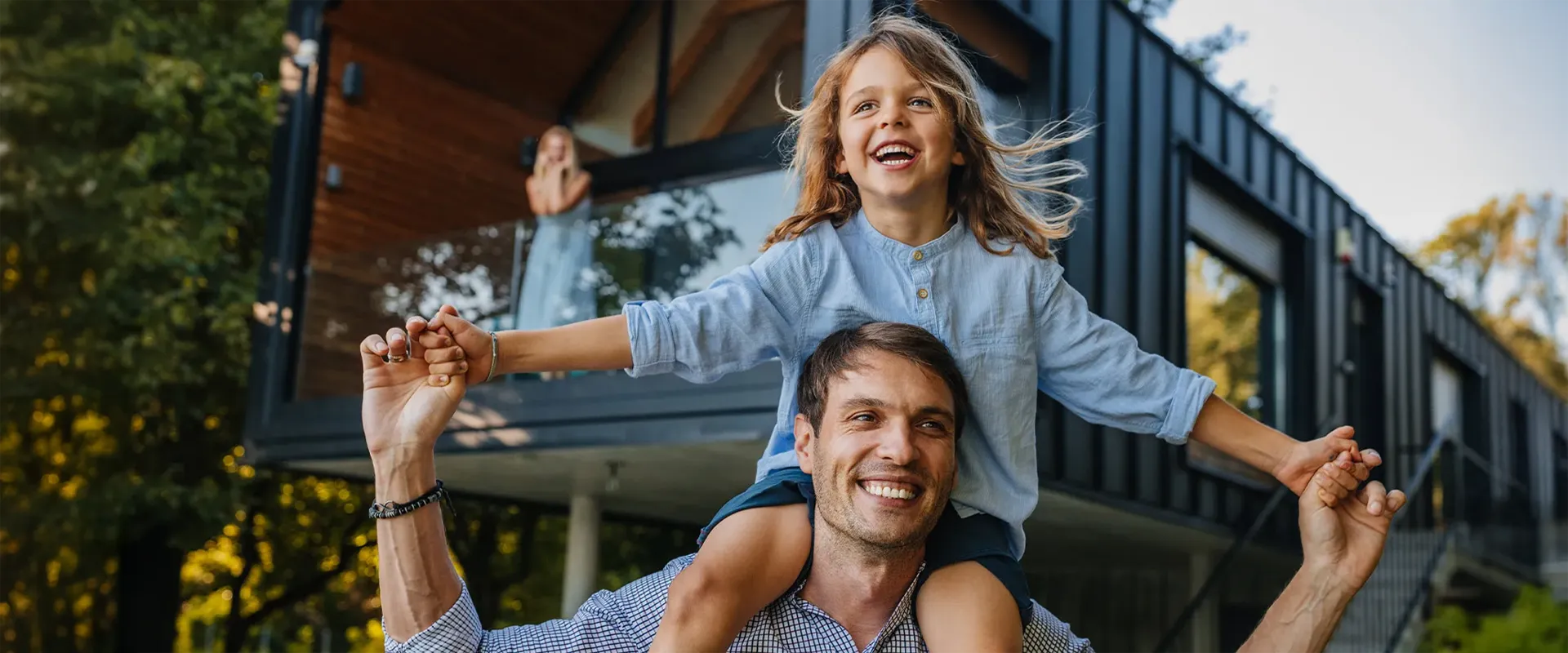 Vater trägt seine lachende Tochter auf den Schultern vor einem modernen Haus, im Hintergrund winkt eine Frau vom Balkon.